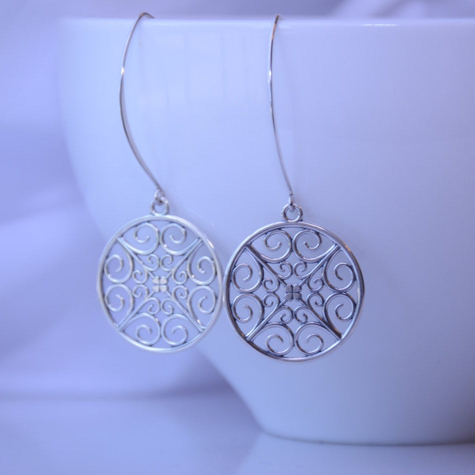 Silver earrings with intricate designs hanging in front of a white bowl.
