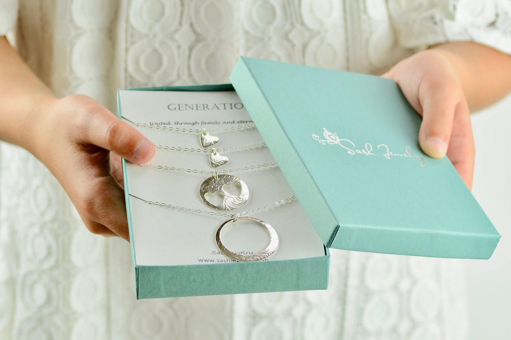 Jewelry set with heart and circle designs in a teal box held by hands against a textured white background.
